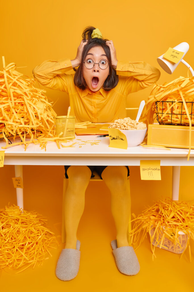 Mujer con gesto de agobio que se lleva las manos a la cabeza, rodeada de notas adhesivas sobre la mesa de su escritorio, con fondo amarillo